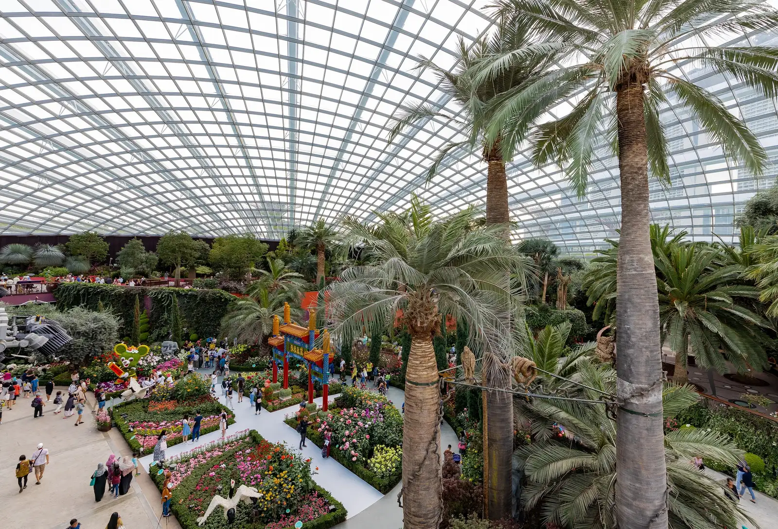 Inside the Flower Dome seen from a raised walkway: tall palms on the right, beds of pink, red and yellow flowers along white paths with visitors, and a festive display with a red-and-gold gateway, all under a vast latticed glass roof