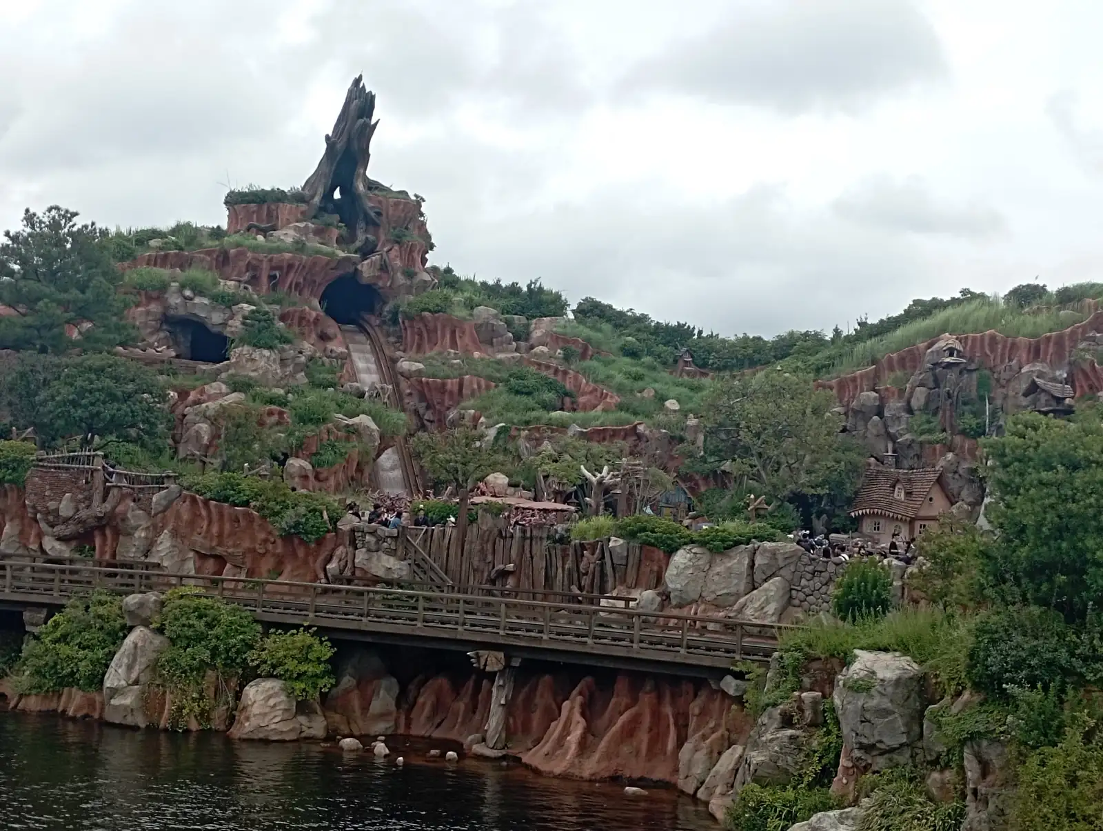 Red-rock mountain covered in grass and pine trees with a log-flume drop and a dead tree on its summit, a wooden bridge and river in front