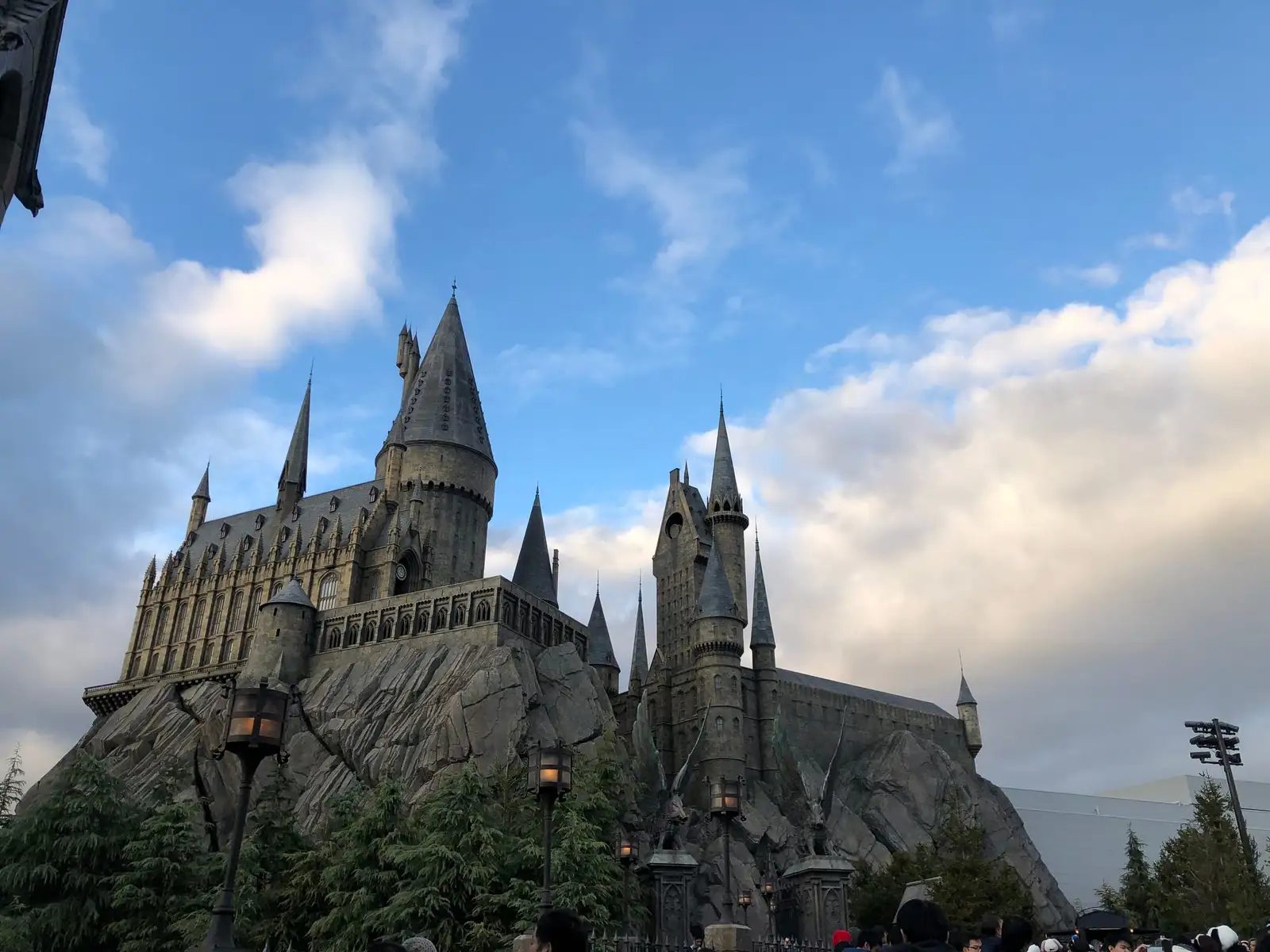 Grey stone Hogwarts castle with tall pointed towers on a rocky hill, under a blue sky with white clouds, lanterns in the foreground