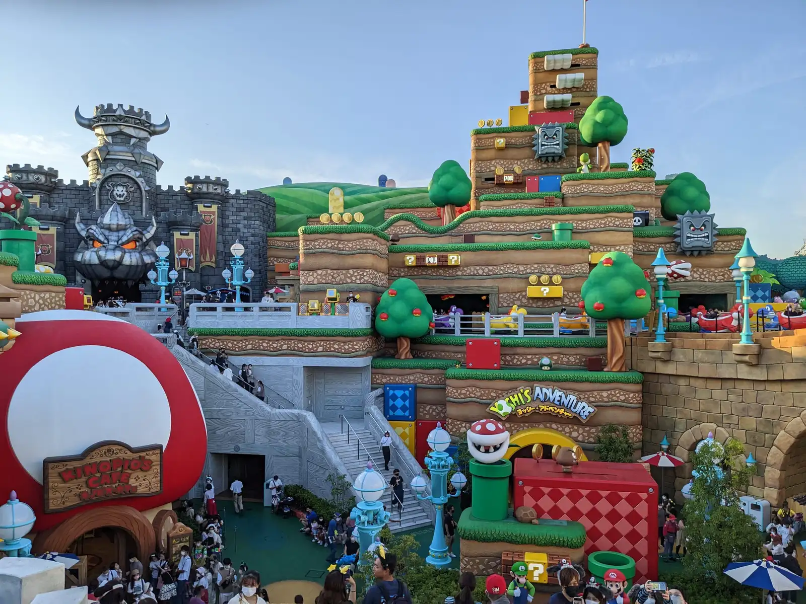 Colourful Super Nintendo World area with Bowser's castle on the left, a giant red mushroom building, green pipes and terraced hills with the Yoshi's Adventure ride