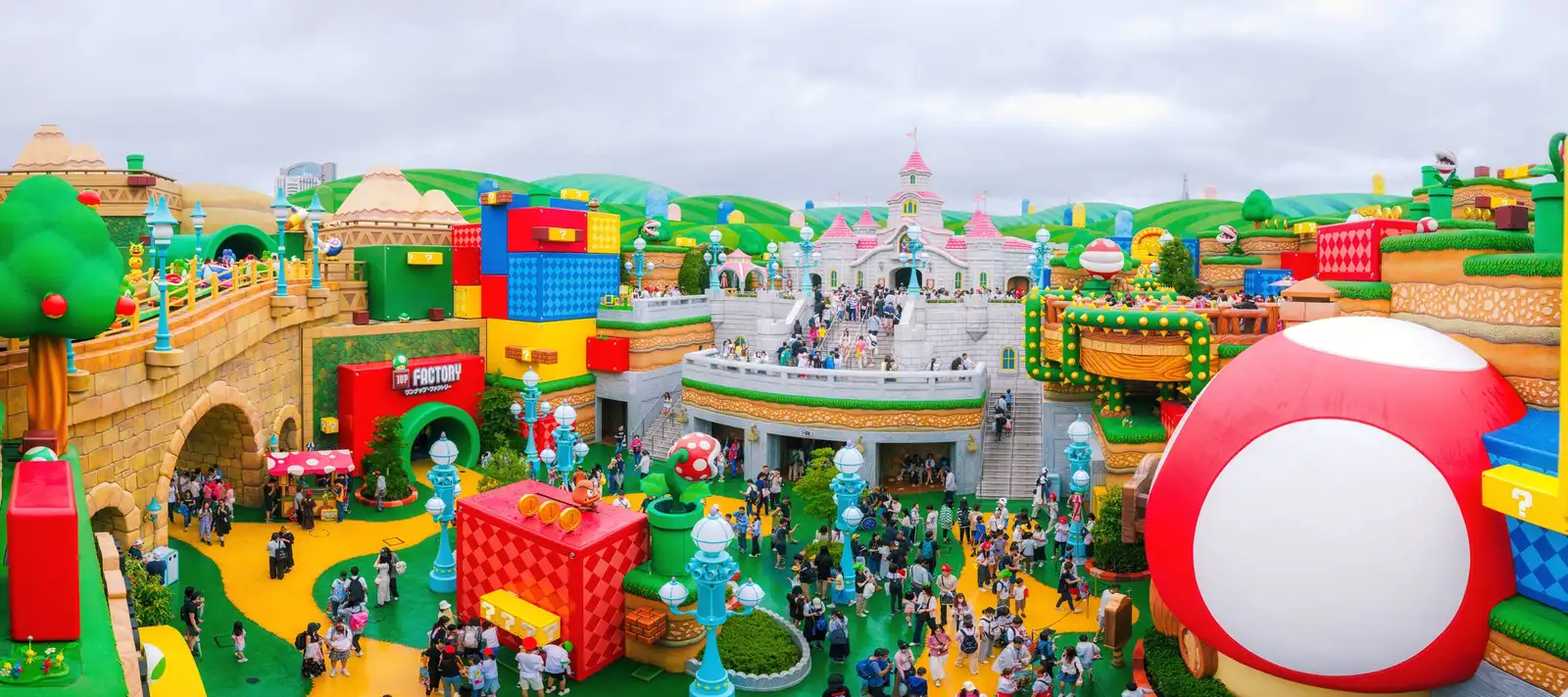 Wide view over Super Nintendo World: Peach's castle on a hill in the centre, giant question blocks and green pipes on the left, a huge red-and-white mushroom building on the right, and crowds on the green and yellow paths below