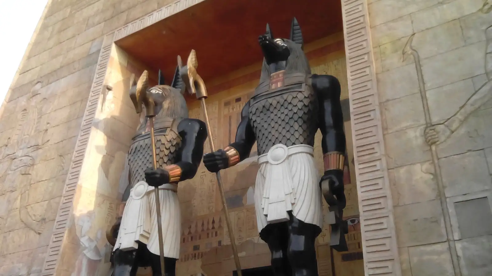 Two giant jackal-headed Anubis statues in black and gold armour with white kilts, holding staffs, guard a doorway in a sandstone wall carved with hieroglyphs